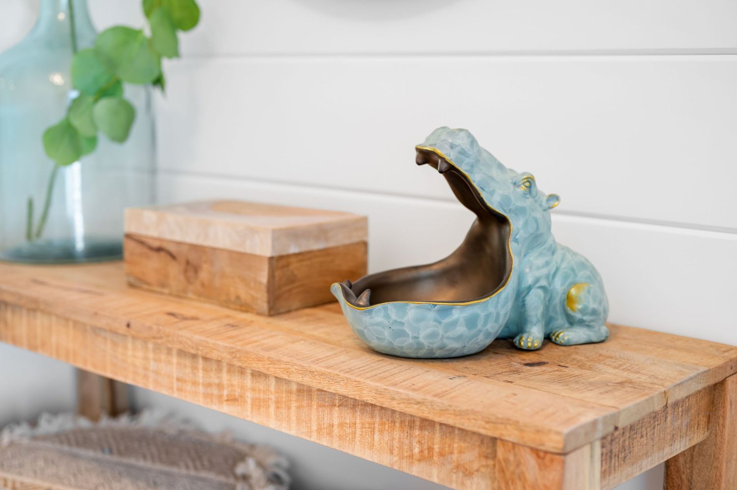 Decorative turquoise hippo bottle opener sits on wooden shelf next to small wooden box and green plant in glass vase.