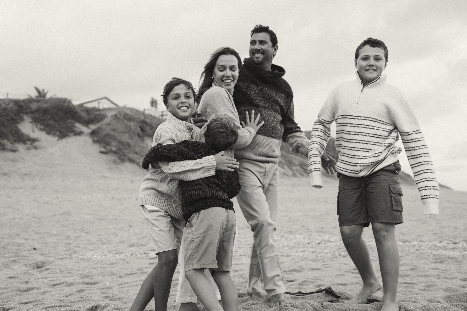A family group laughing and embracing while standing barefoot on a beach.