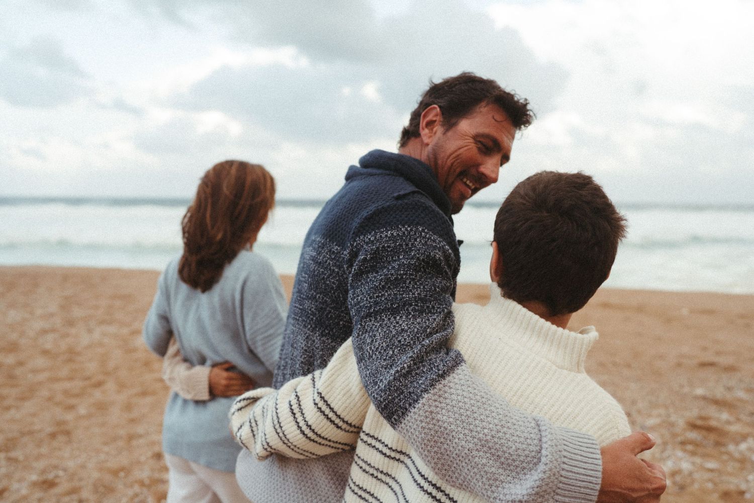 Candid beach moment showing people in warm sweaters embracing by the ocean.