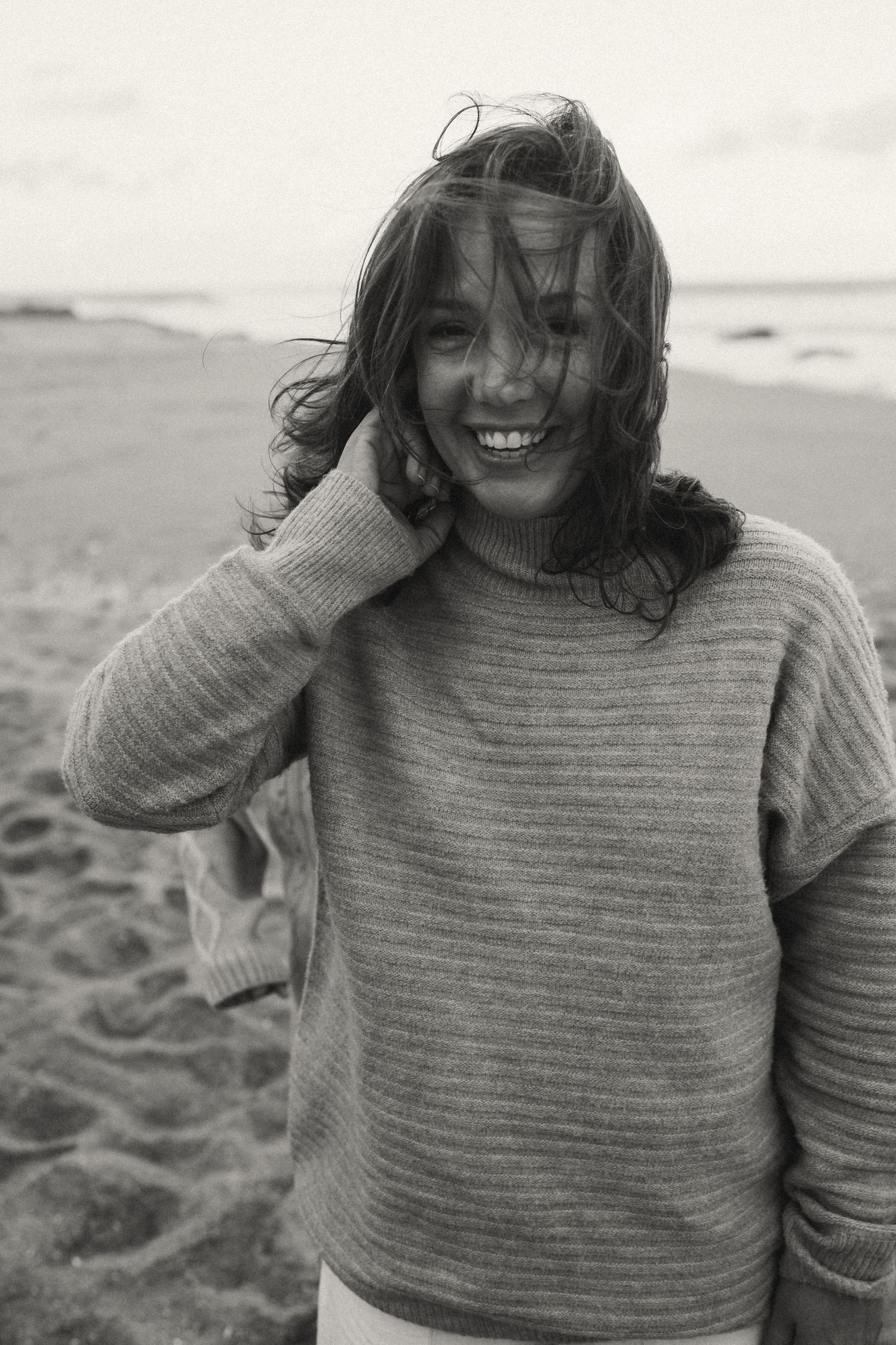 Person in a striped sweater laughing with windblown hair on the beach.