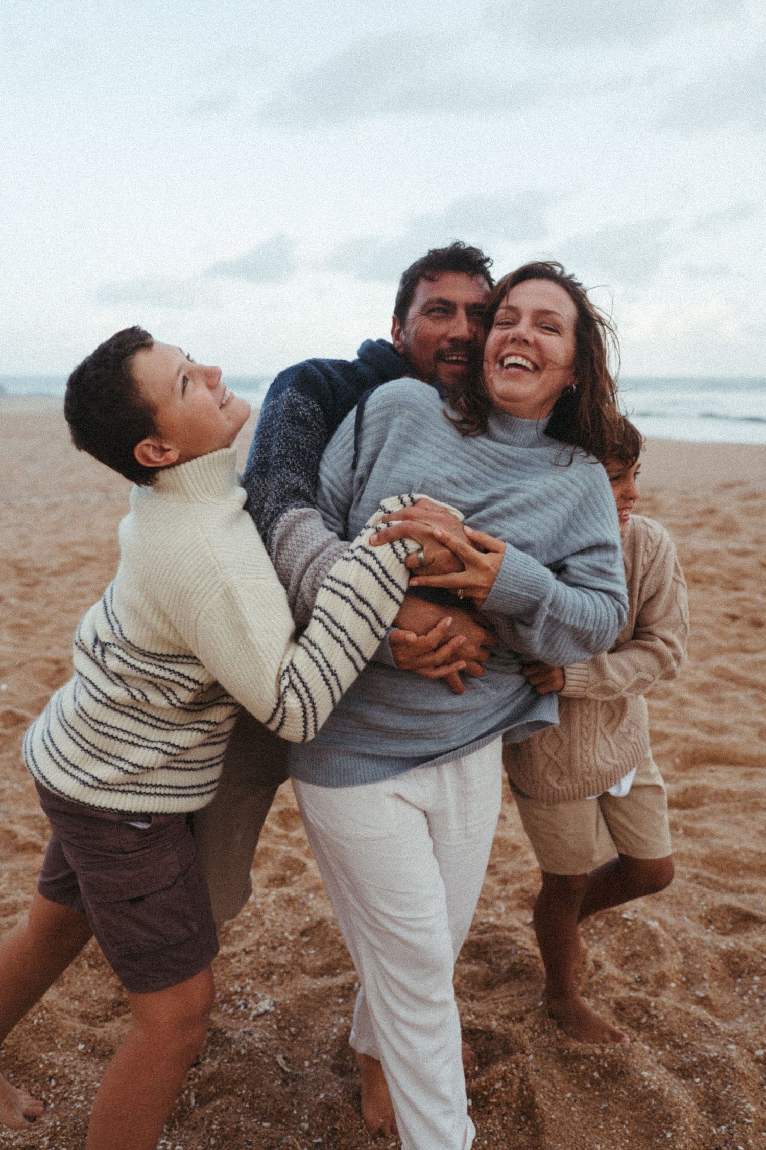 Three friends laughing and embracing together on a beach at sunset wearing cozy sweaters.