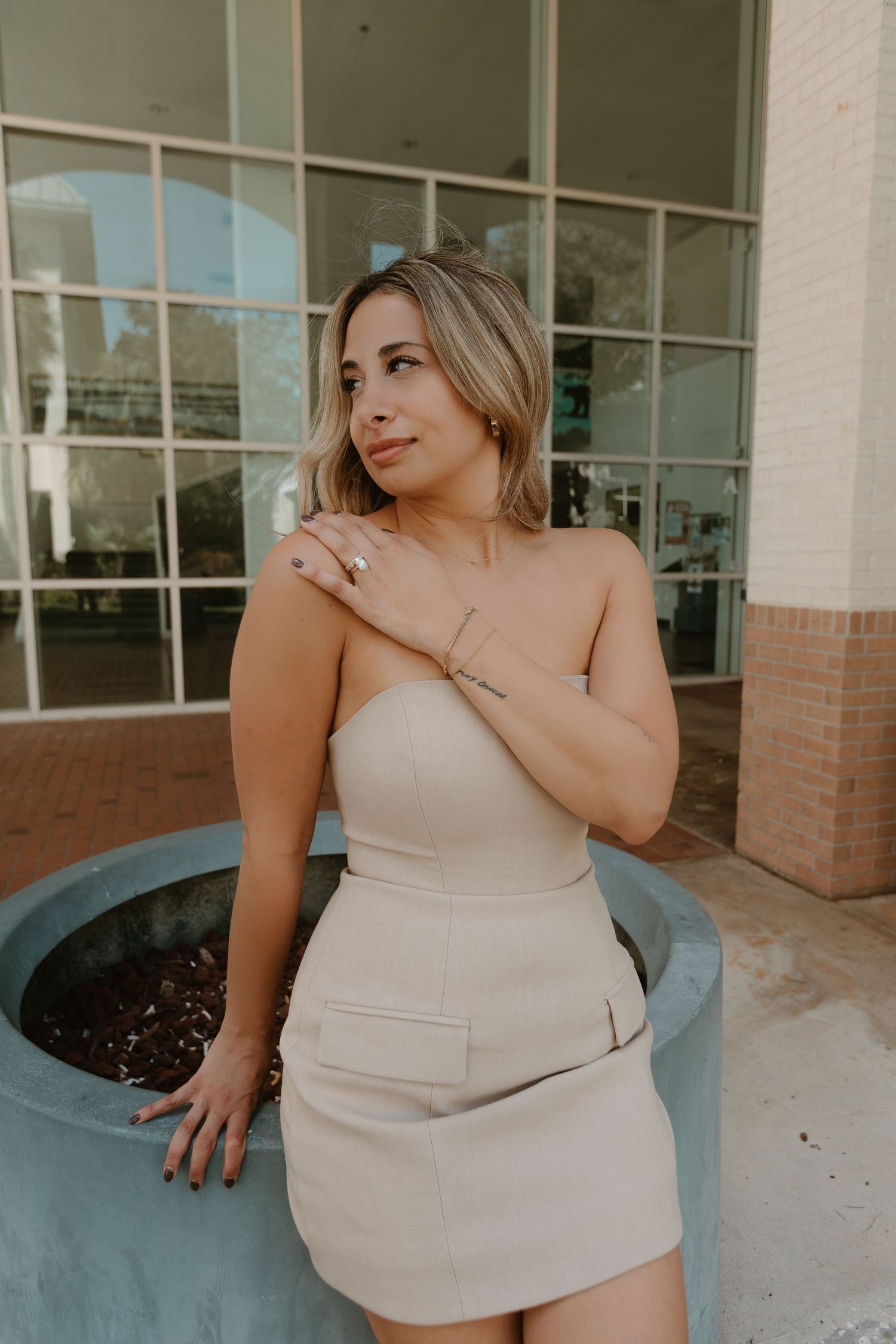 Woman in beige dress poses casually outside brick building with glass windows.