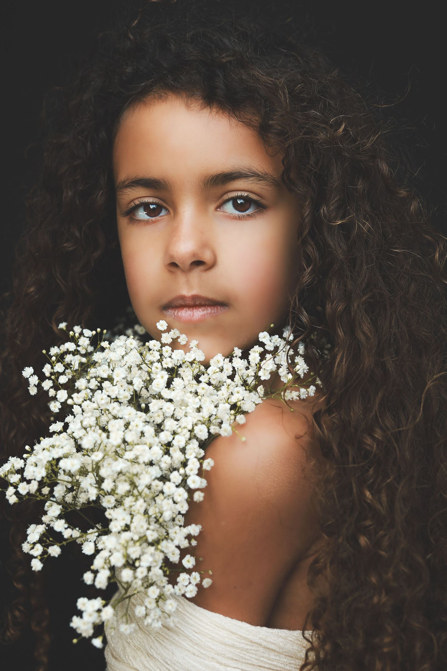 Young girl with curly hair holding white baby's breath flowers against a dark background.