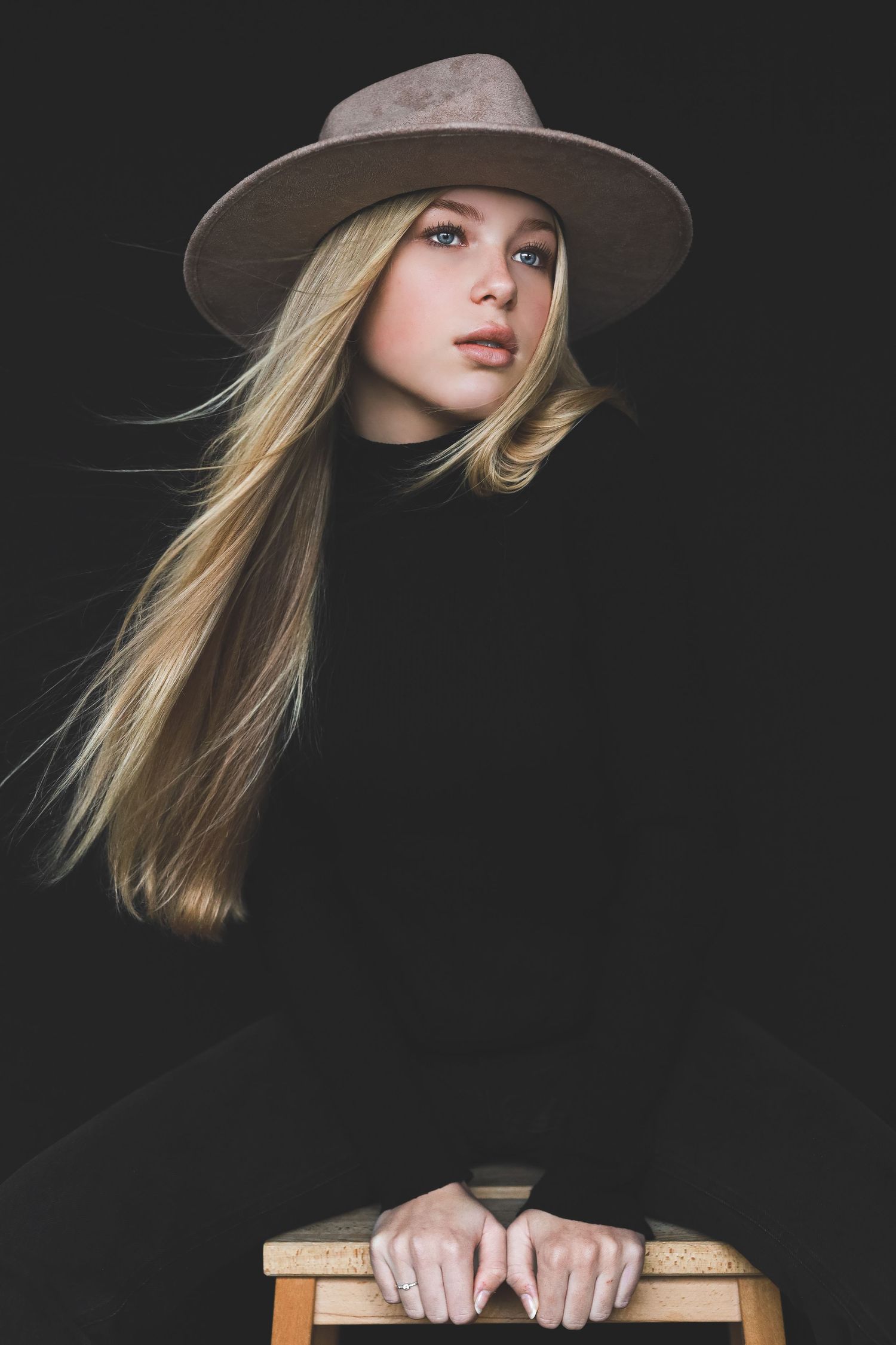 Young blonde woman in a wide-brim hat and black turtleneck poses on a wooden stool against a dark background.