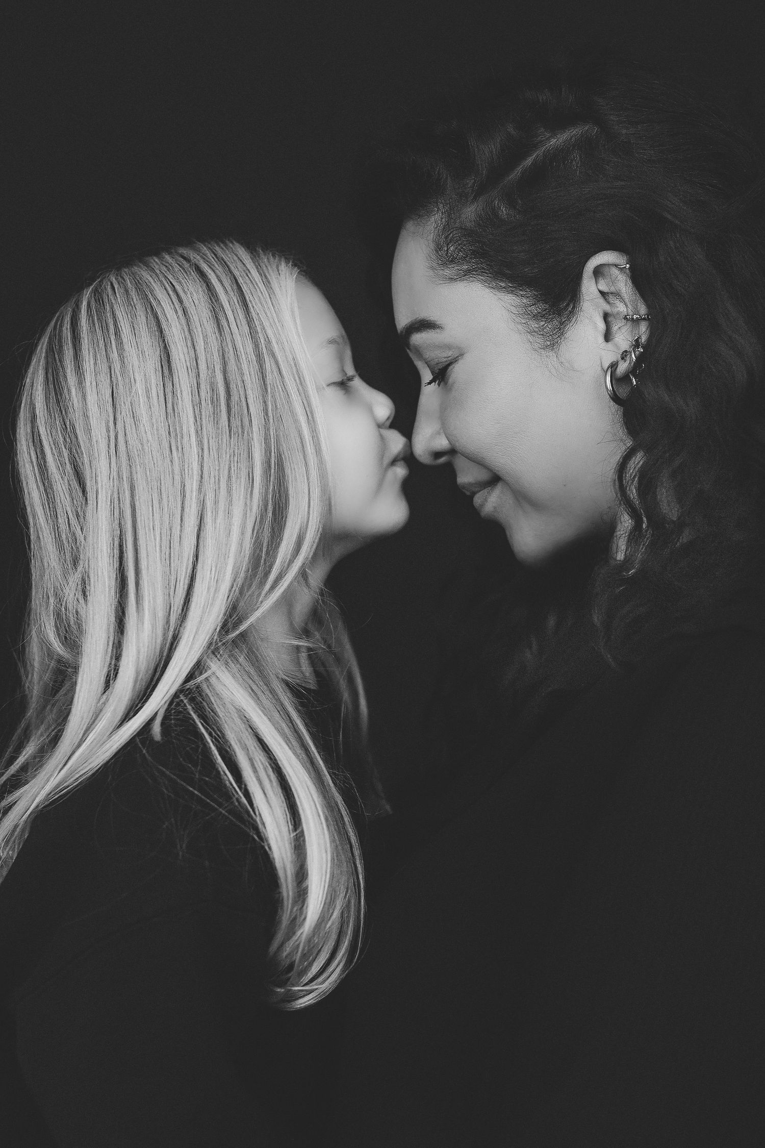 Black and white portrait of a mother and young blonde daughter touching noses, sharing a tender moment against dark background.