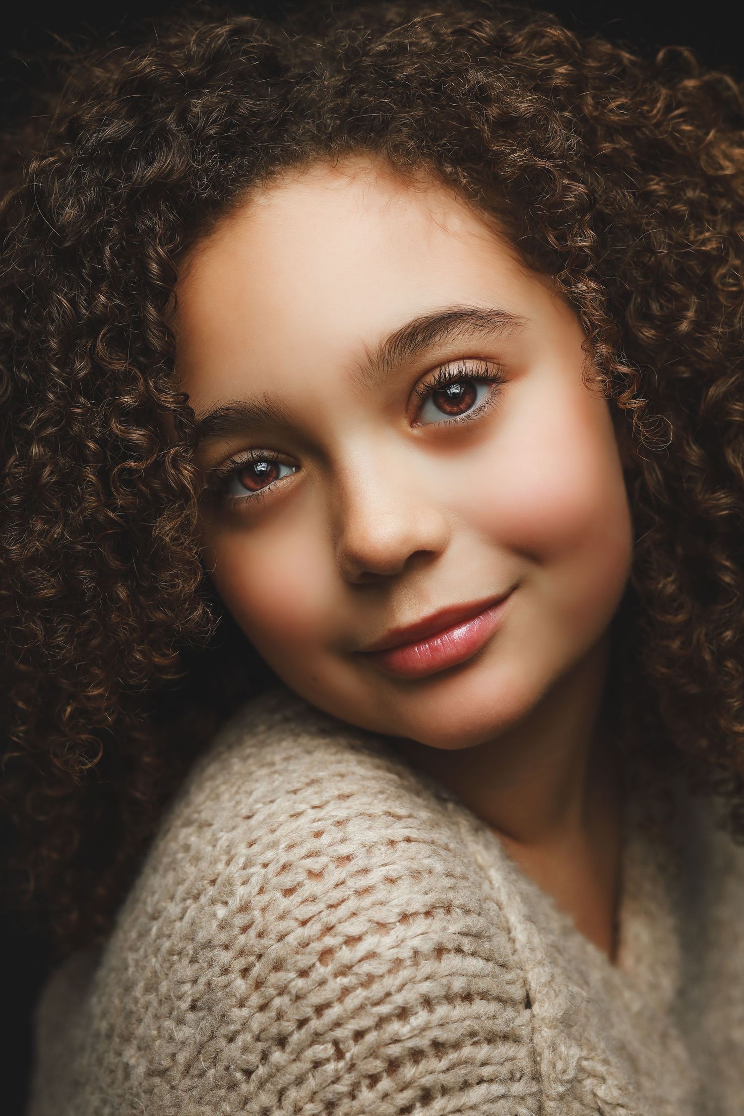 Young girl with curly brown hair and warm smile wearing a cozy knit sweater against dark background.