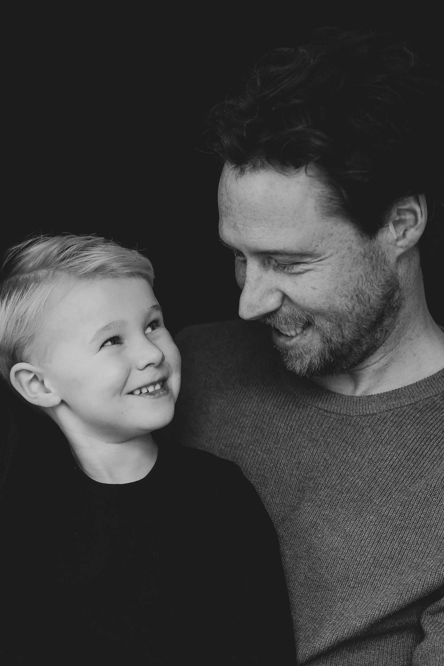 Black and white portrait of a smiling young boy looking up at his bearded father against dark background.