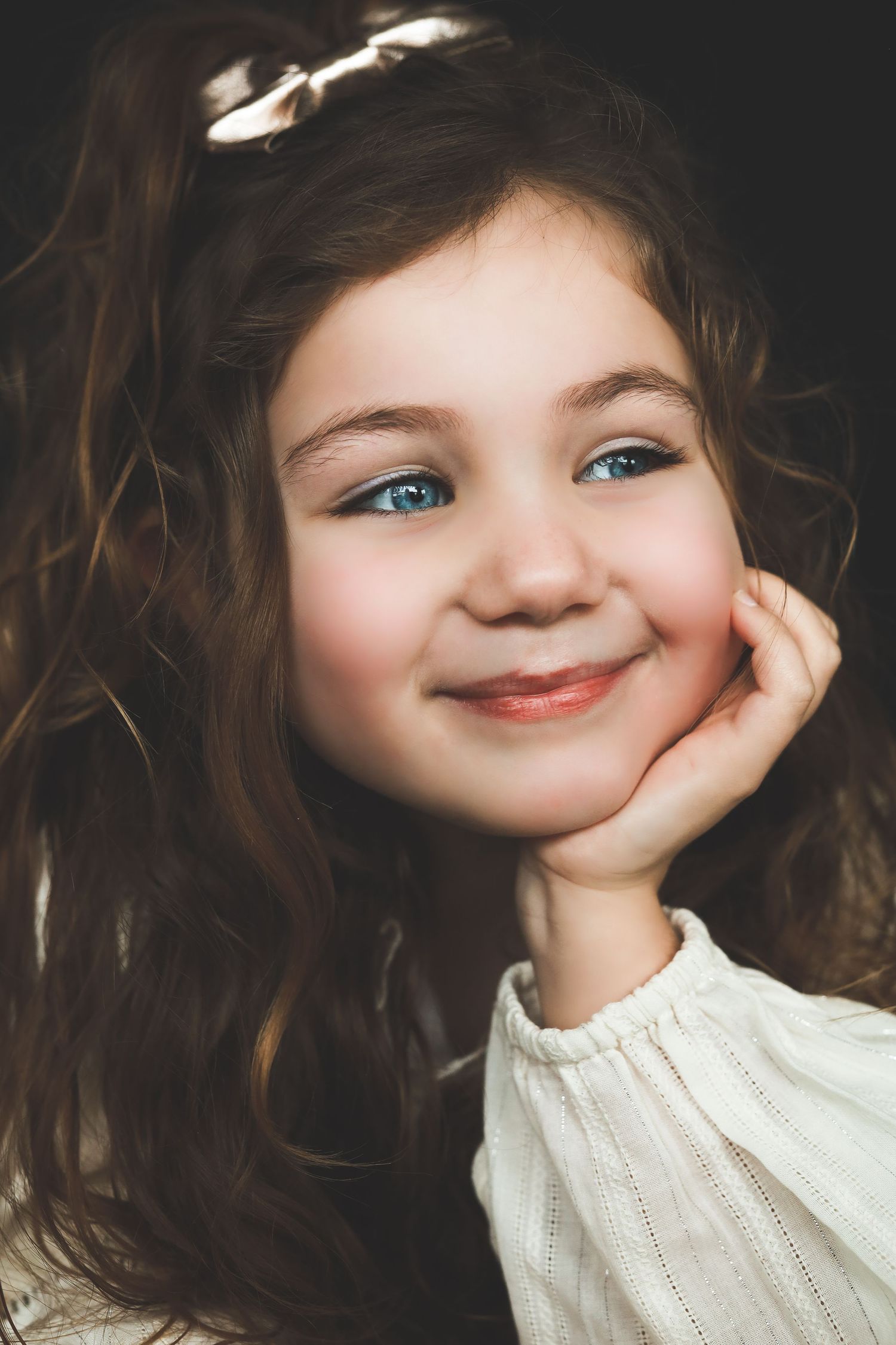 A smiling young girl with curly brown hair and blue eyes rests her chin on her hand, wearing a white dress.