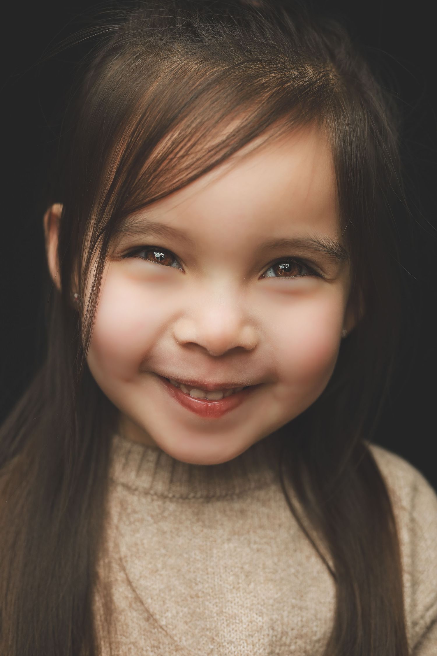 A smiling young girl with long brown hair wearing a beige turtleneck sweater against a dark background.