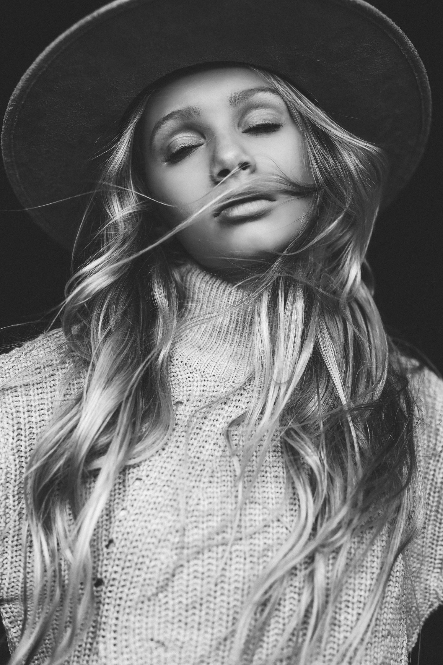 Black and white portrait of a blonde woman in a wide-brim hat and knit sweater with hair blowing across her face.