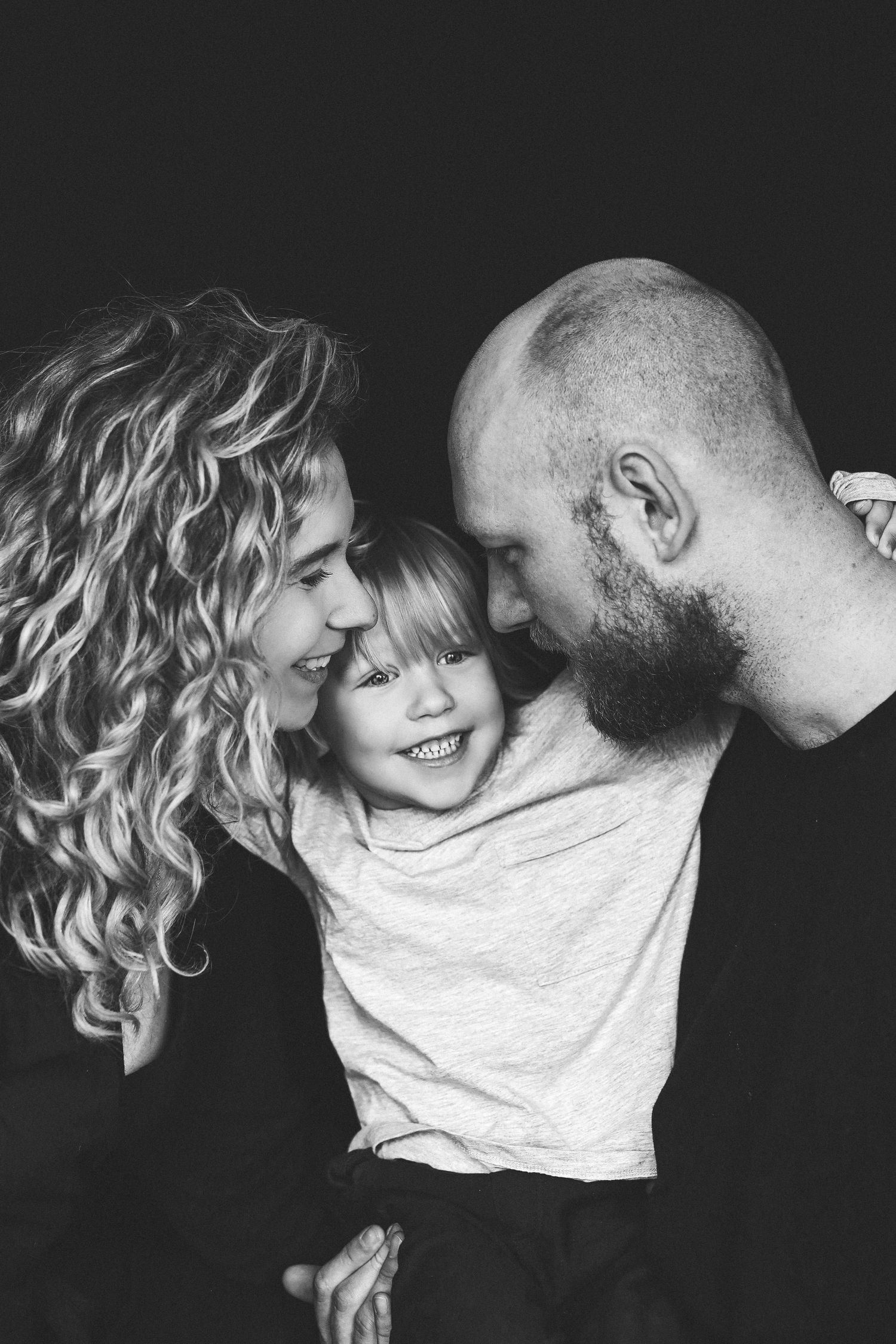 Black and white family portrait of smiling parents with curly and bald hair embracing their happy laughing young child.