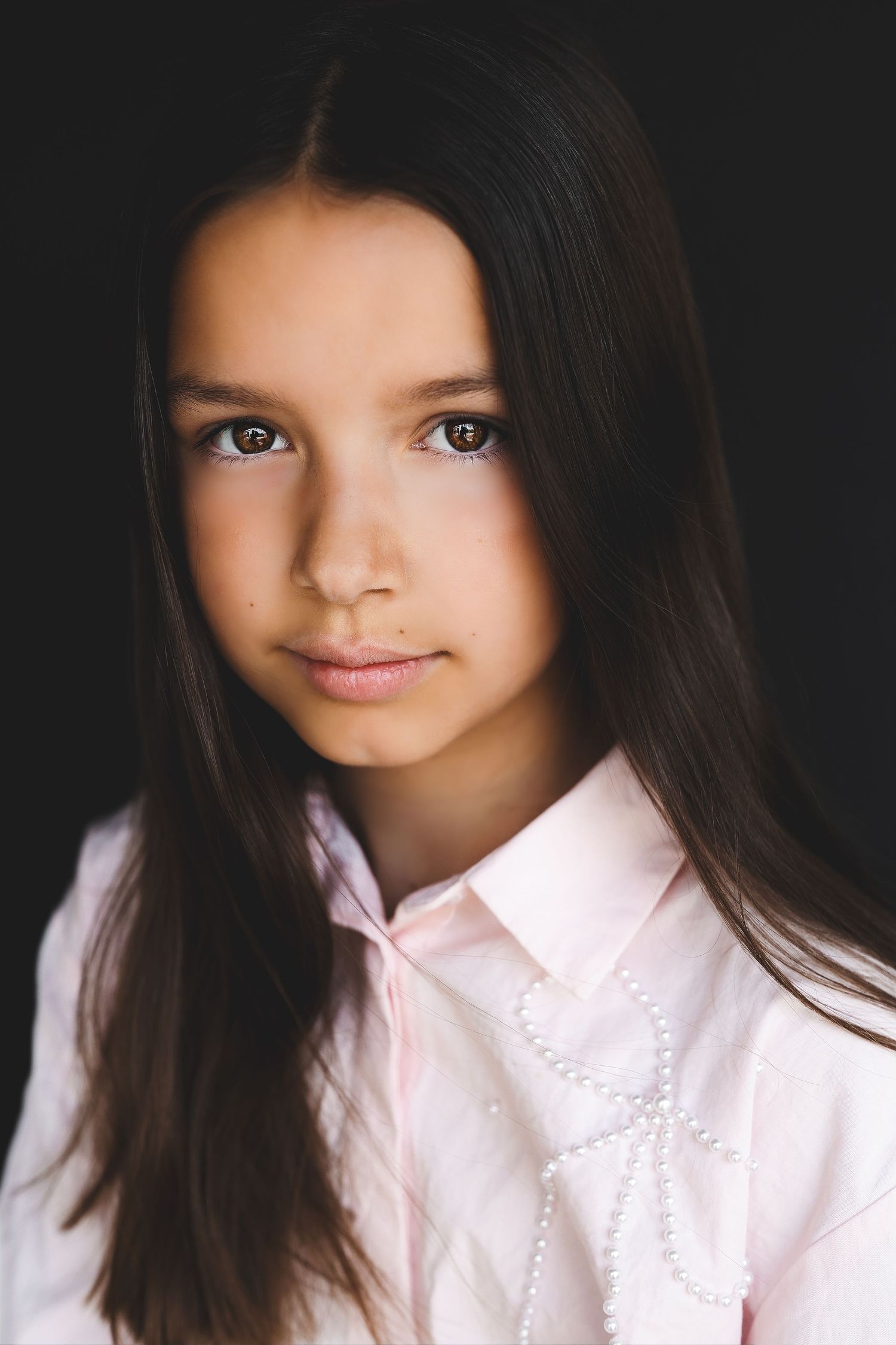 Young girl with long dark hair wearing a pink pearl-embellished blouse, posing against a dark background.
