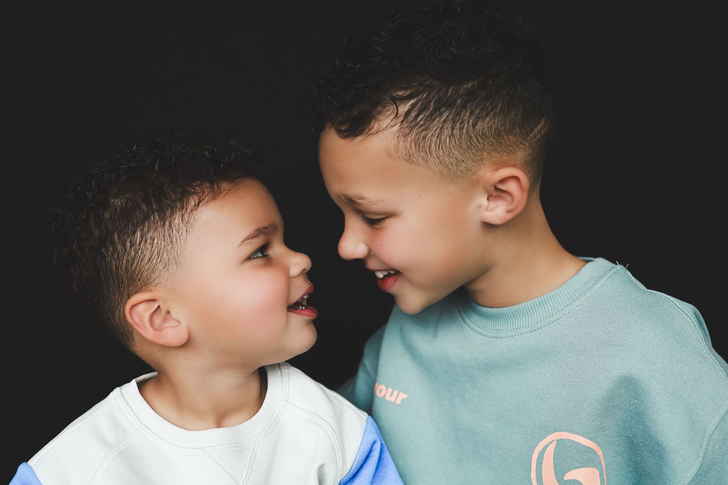 Two young boys smiling and facing each other against a dark background, sharing a joyful sibling moment.