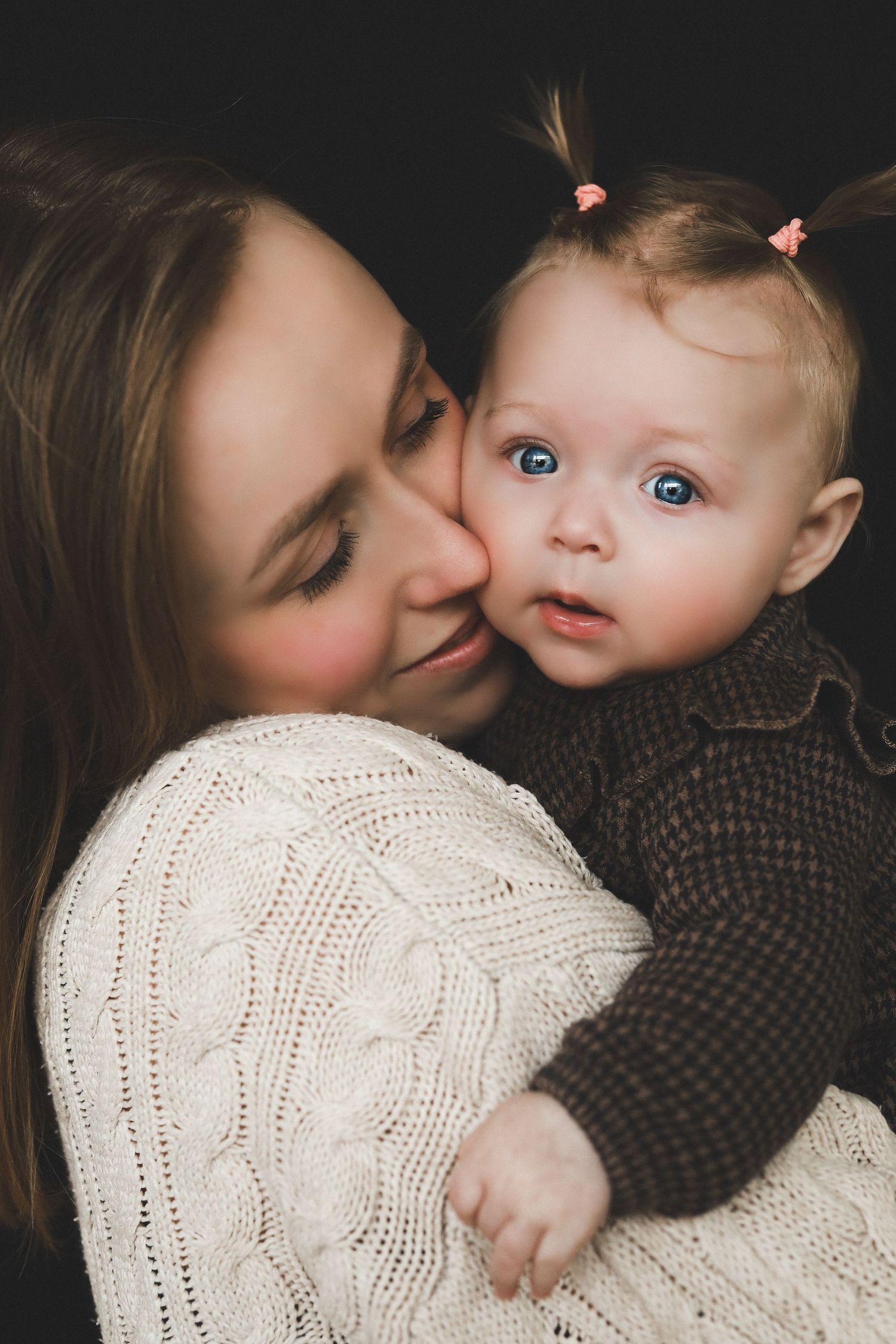 A young mother in a white knit sweater lovingly kisses her blue-eyed baby girl with tiny pigtails.