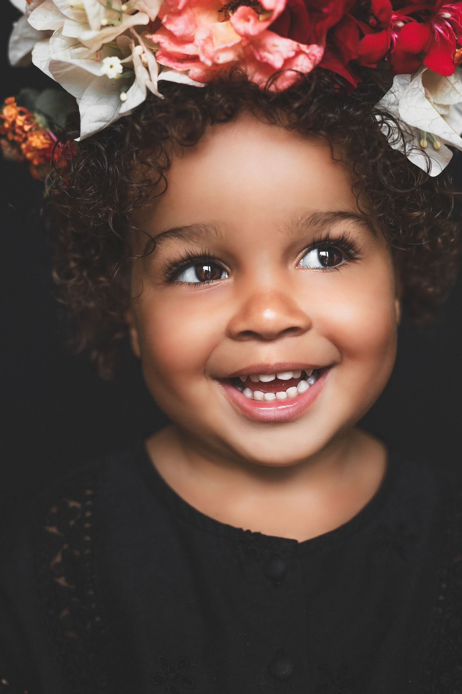 Smiling toddler girl with curly hair wearing a colorful floral crown, dressed in black against a dark background.