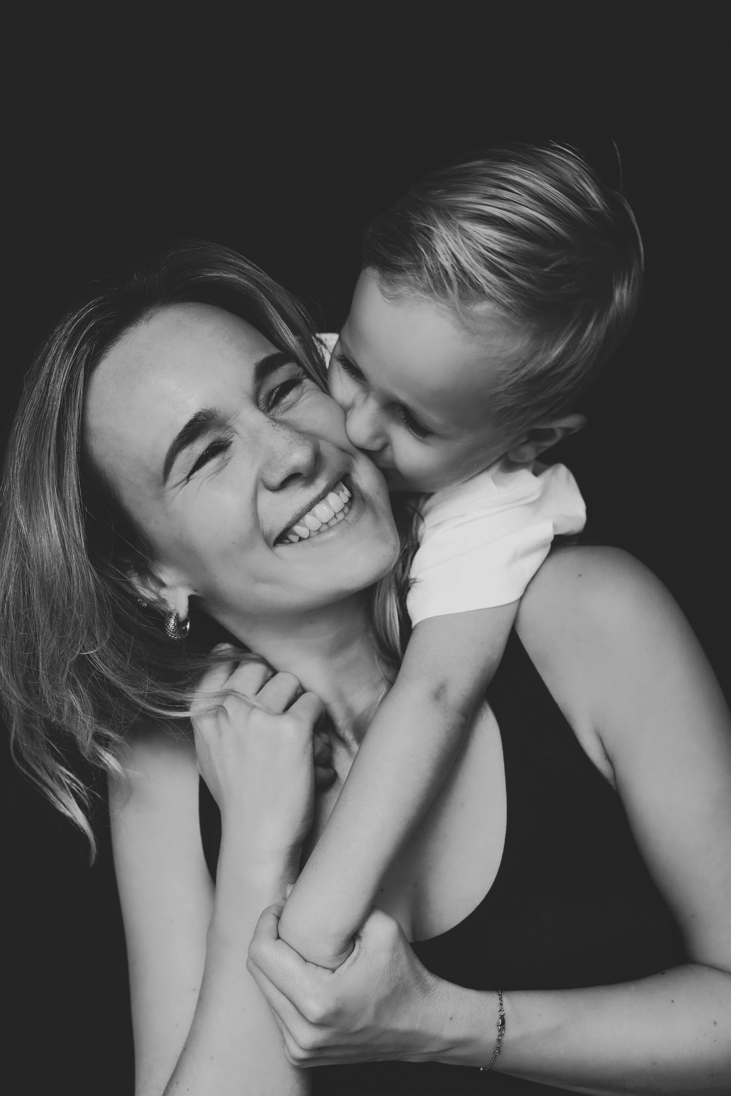 Black and white photo of a smiling woman being kissed on the cheek by a young child in a tender moment.