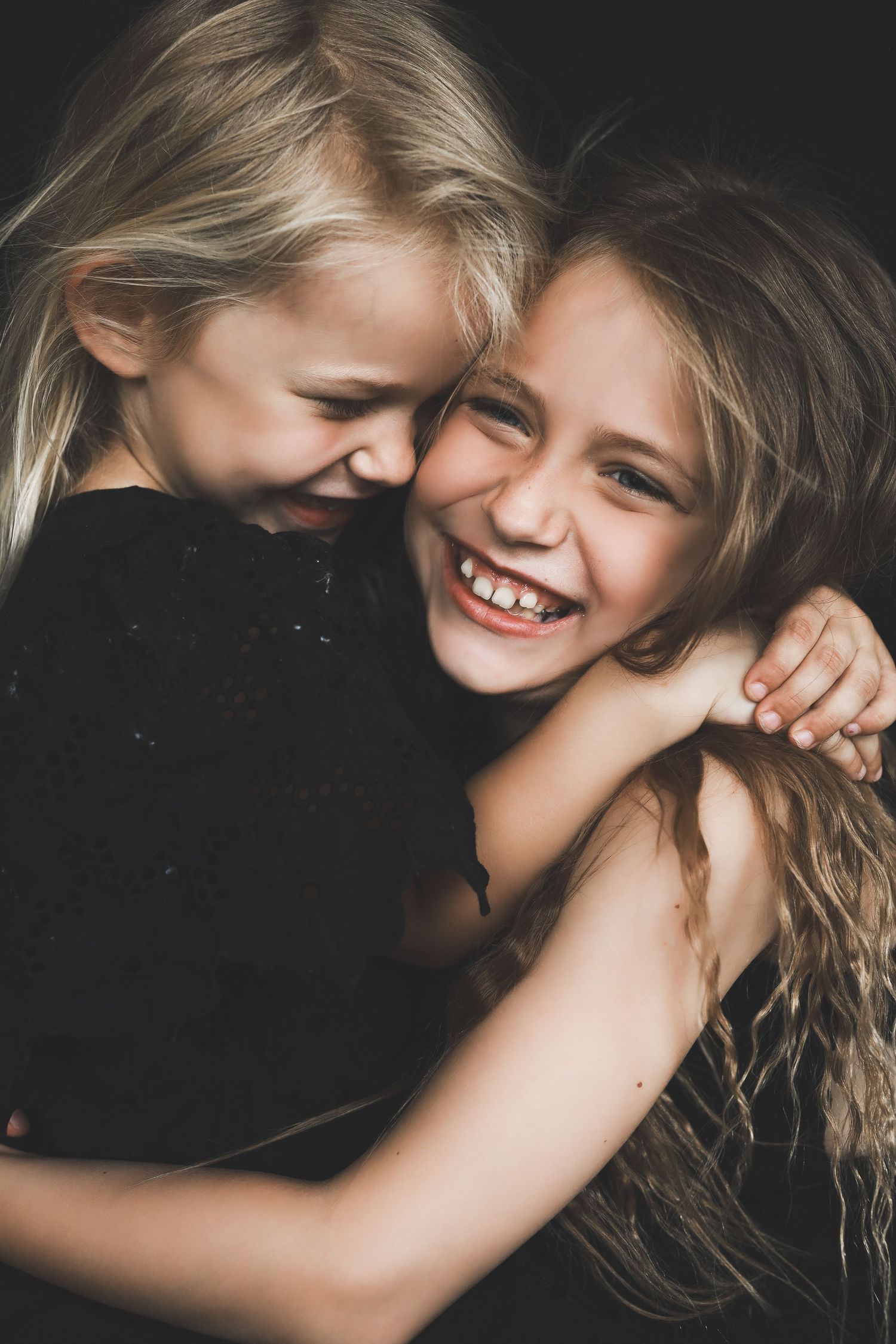 Two young blonde girls in black outfits laughing and hugging each other against a dark background.