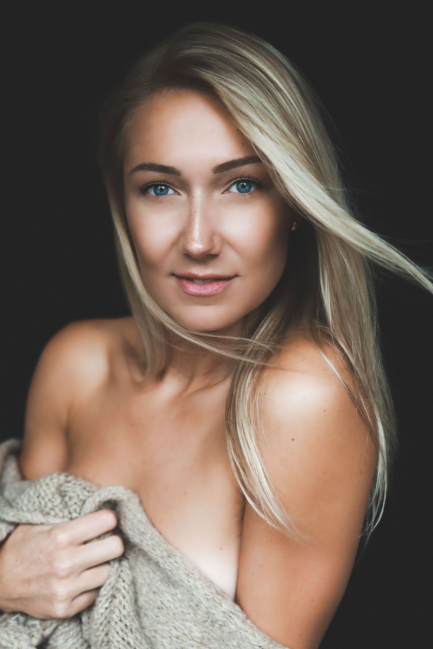 Blonde woman with blue eyes wrapped in a cozy knit sweater posing against a dark background.