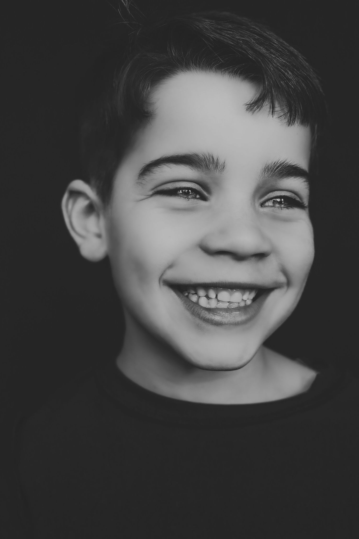 Black and white portrait of a smiling young boy with bright eyes against a dark background.