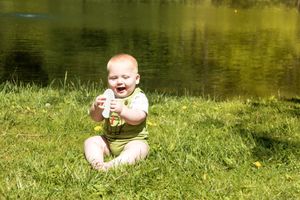 A baby in green overalls sits smiling on grass near a pond on a sunny day.