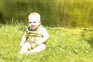 A baby in light green clothing sits on grass with a pond reflecting golden light in the background.