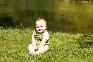 A small baby sits on grass by a serene pond on a bright sunny day.