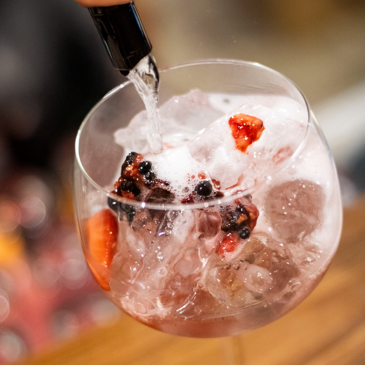 Bartender pouring tonic water into a berry gin and tonic cocktail over ice in balloon glass.