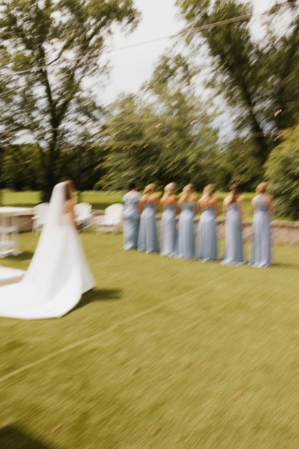 Blurred motion shot of bridesmaids in light blue dresses standing in line on a green lawn.