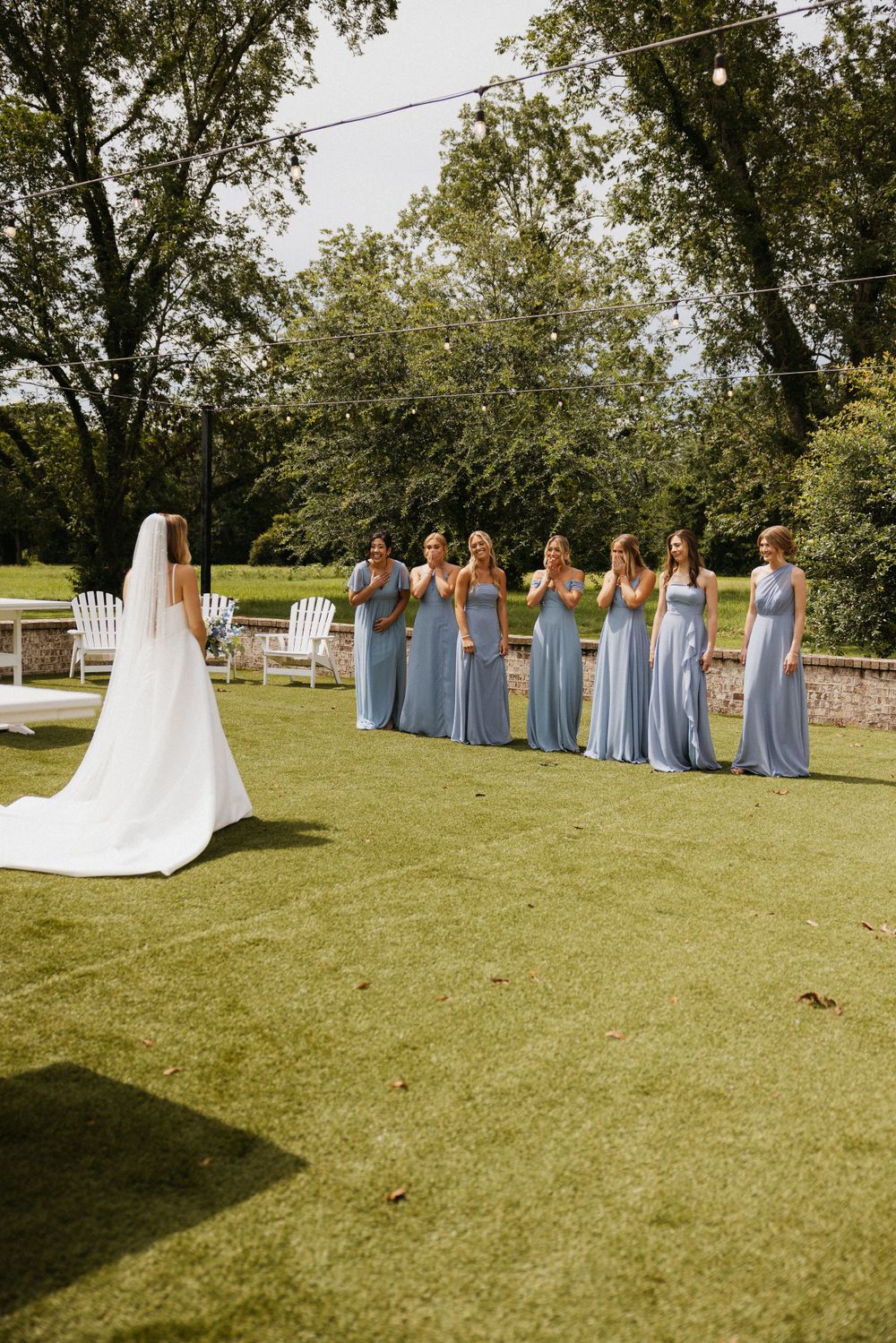 Bridesmaids in powder blue dresses stand in line outdoors while facing the bride in her white gown.