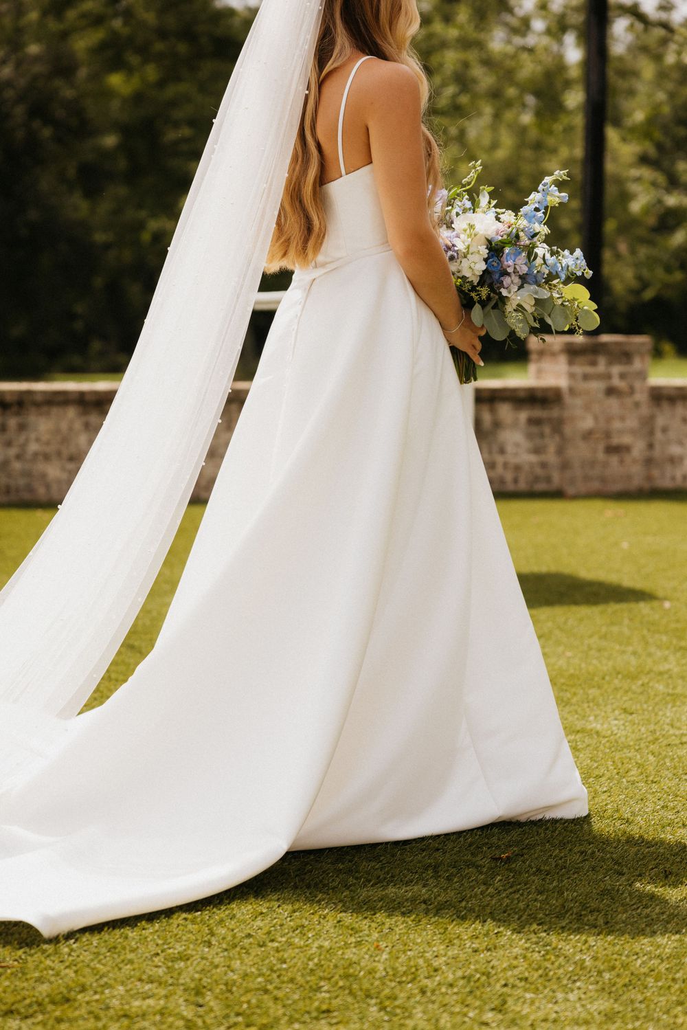 Back view of wedding dress featuring delicate straps and flowing train with white and blue bouquet.