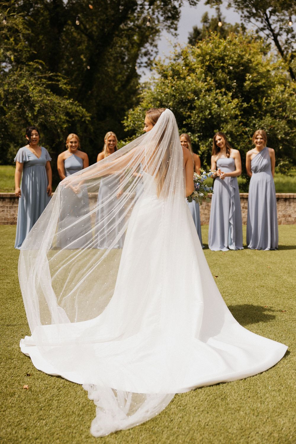 Bride in flowing white gown and veil spins on green lawn as bridesmaids watch in powder blue dresses.
