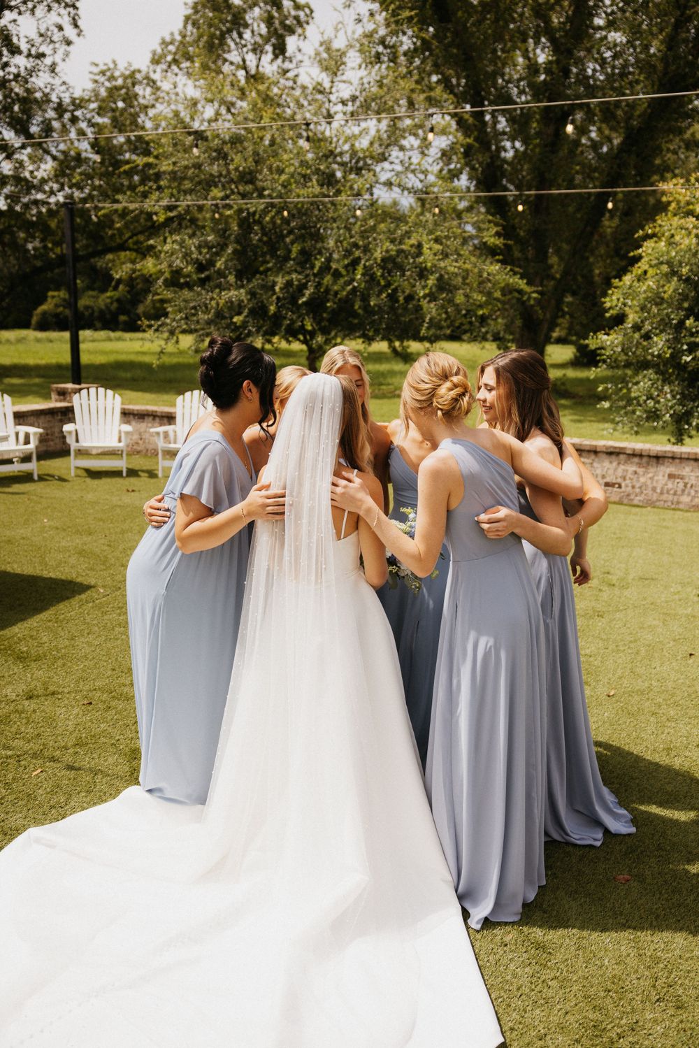 Intimate moment as bridesmaids in blue dresses gather around the bride on a sunny lawn.