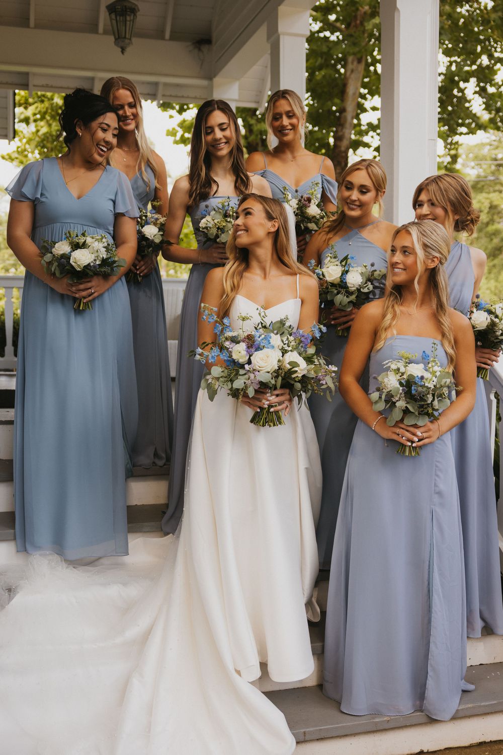 A bridal party wearing dusty blue bridesmaid dresses and white wedding gown pose together on a porch with matching bouquets.