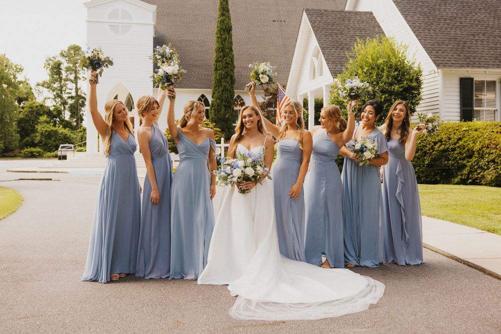 A wedding party stands outdoors in coordinating light blue dresses holding floral bouquets.