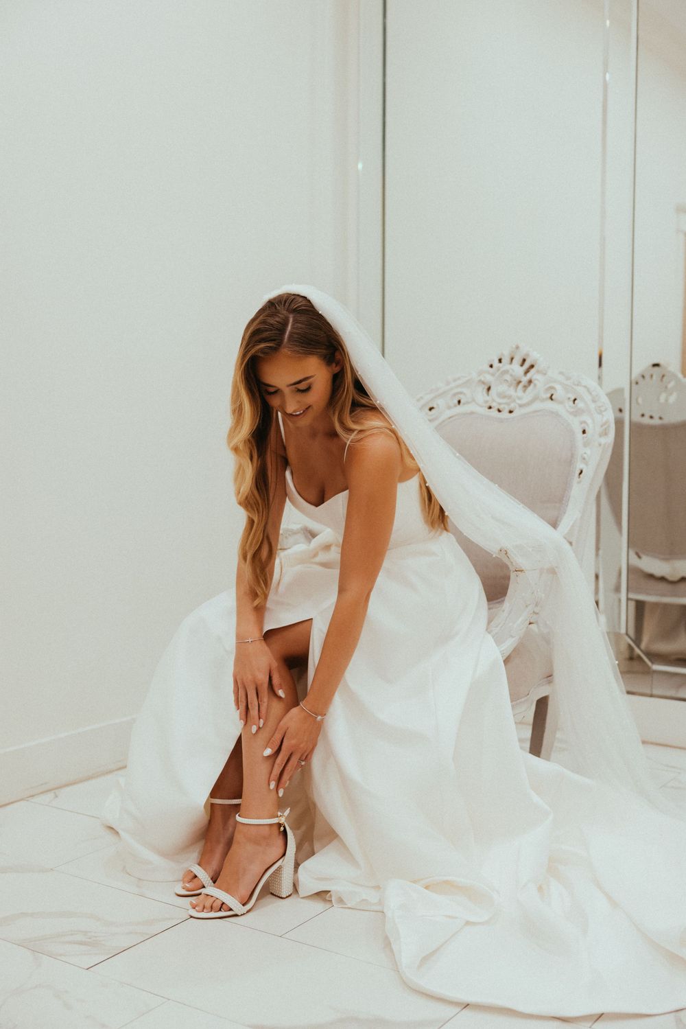 Bride in white wedding dress adjusting her shoe while sitting on vintage chair.
