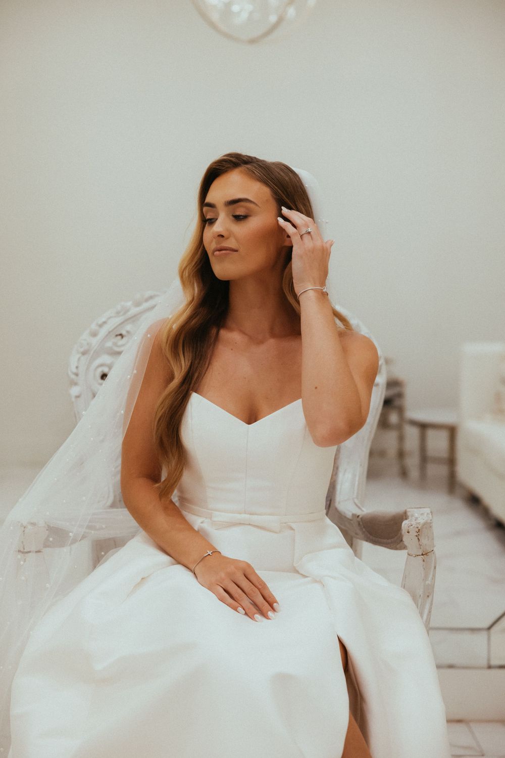 Elegant bride in a classic white strapless wedding gown sits gracefully in a vintage-style chair.
