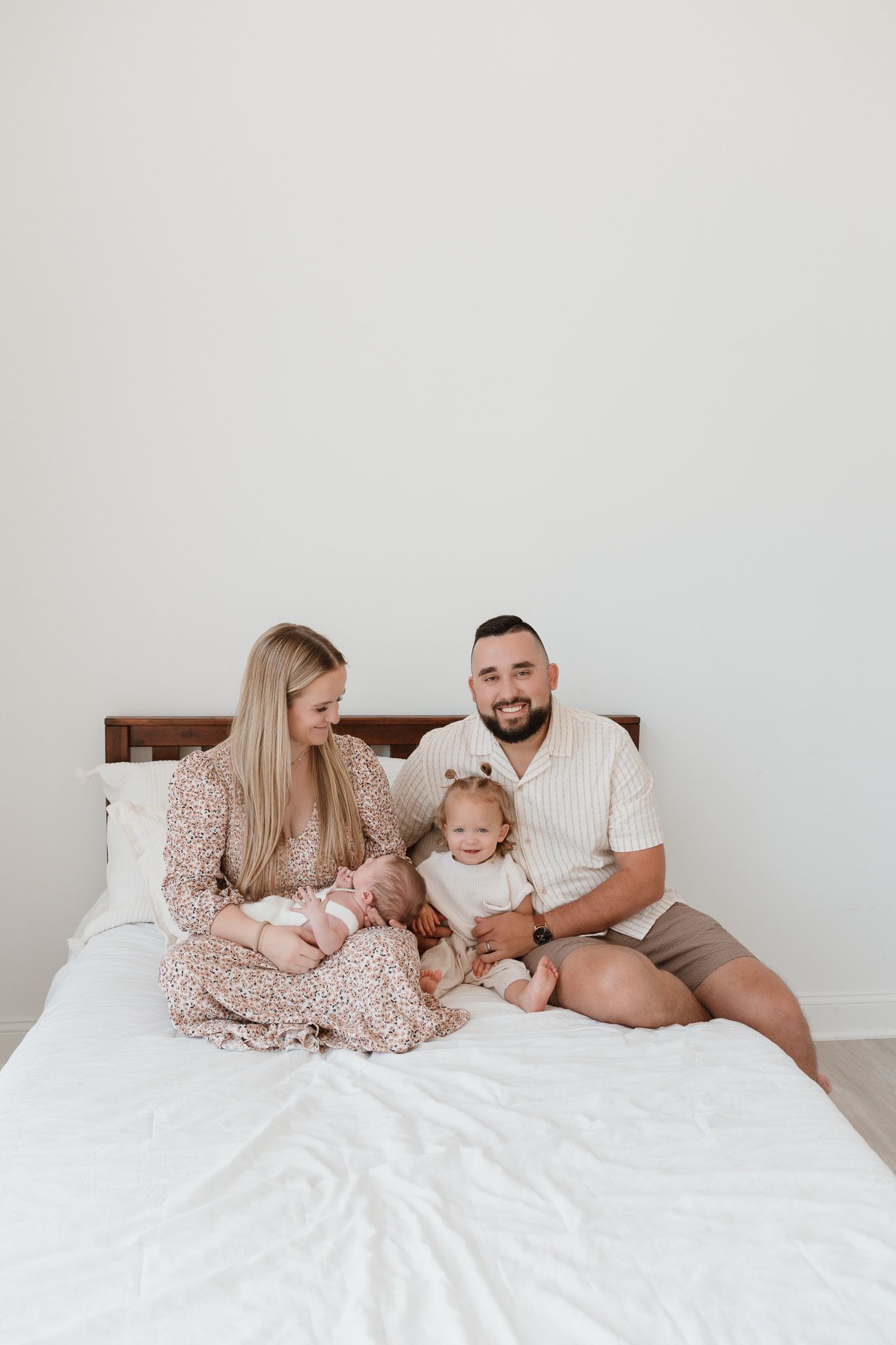 Family seated on a bed in a minimalist room, the father is smiling at the camera while the mother is smiling at her children.