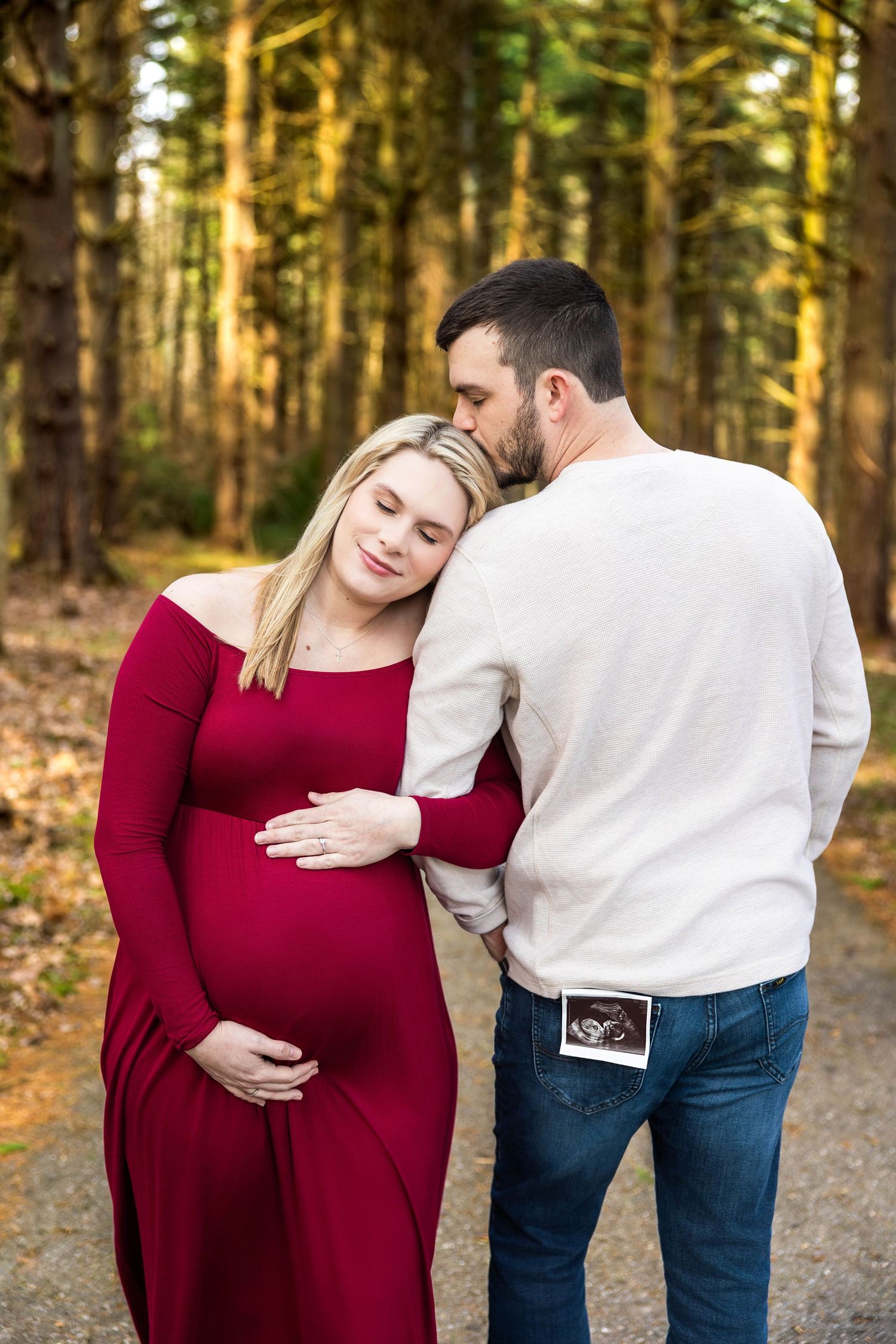 A couple shares an intimate moment in a forest setting while holding a sonogram photo.