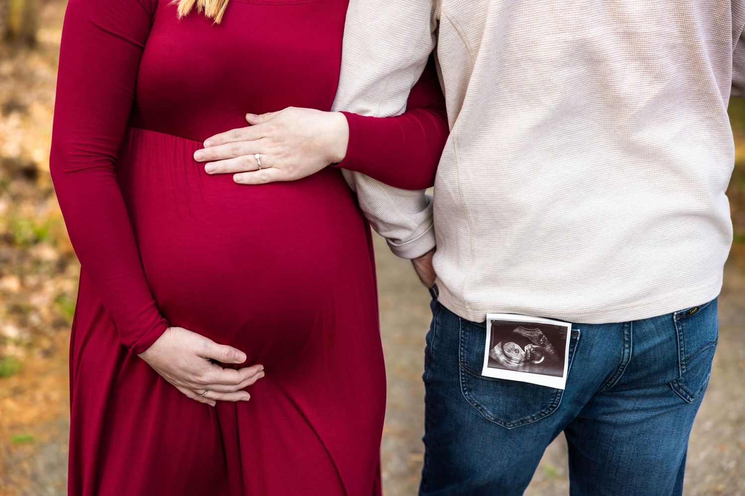 Couple in autumn setting with burgundy maternity dress holding ultrasound photo in back pocket of jeans.