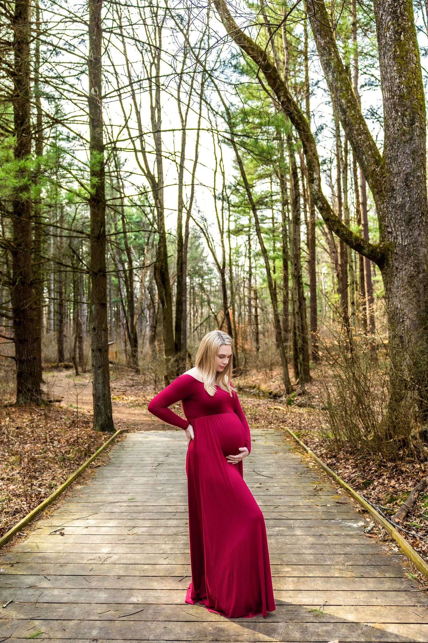 A figure in a flowing burgundy maternity dress stands on a wooden boardwalk path through an autumn forest.