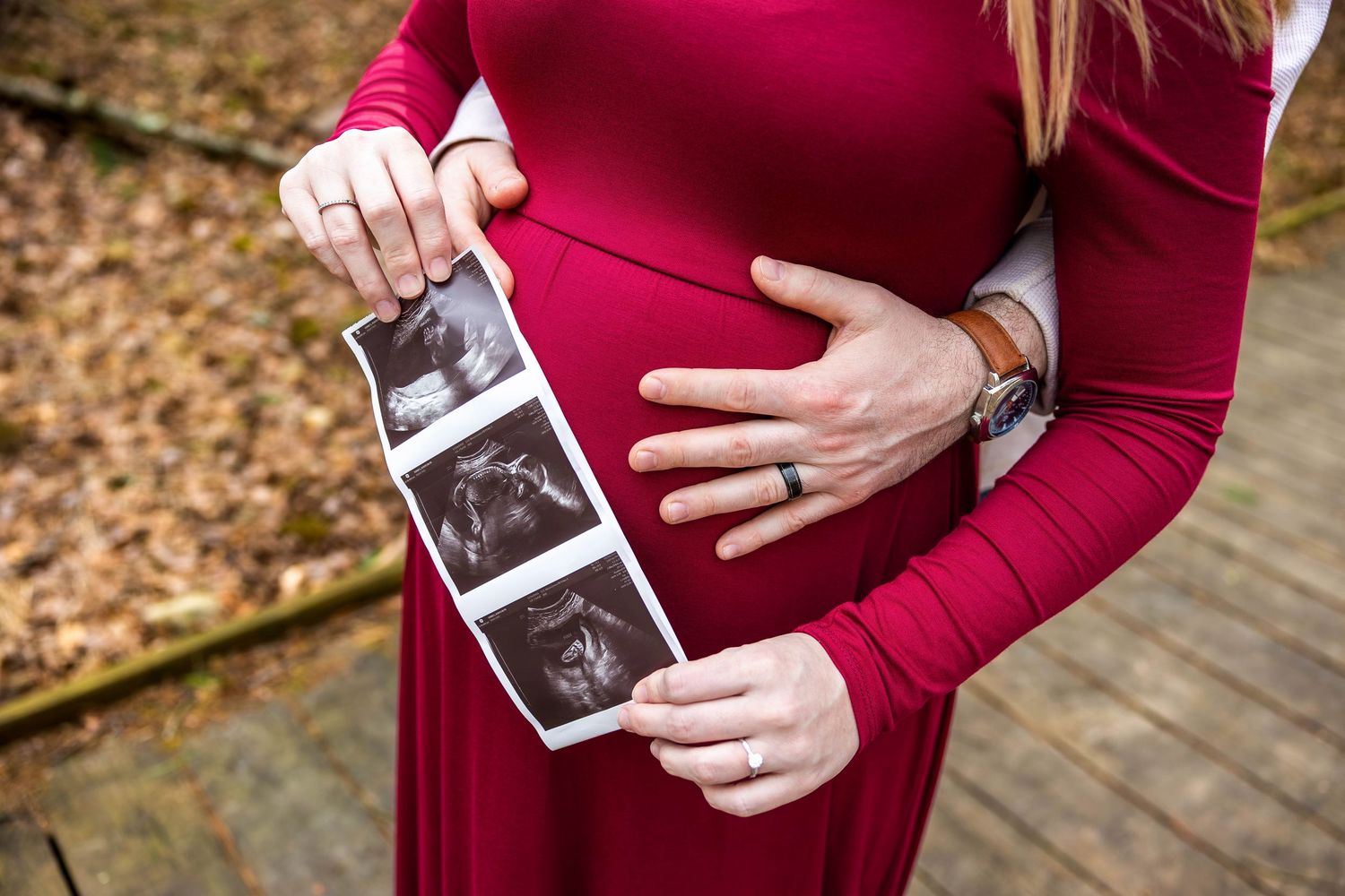A person in a burgundy sweater holds a sonogram photo while cradling their pregnant belly in an autumn setting.