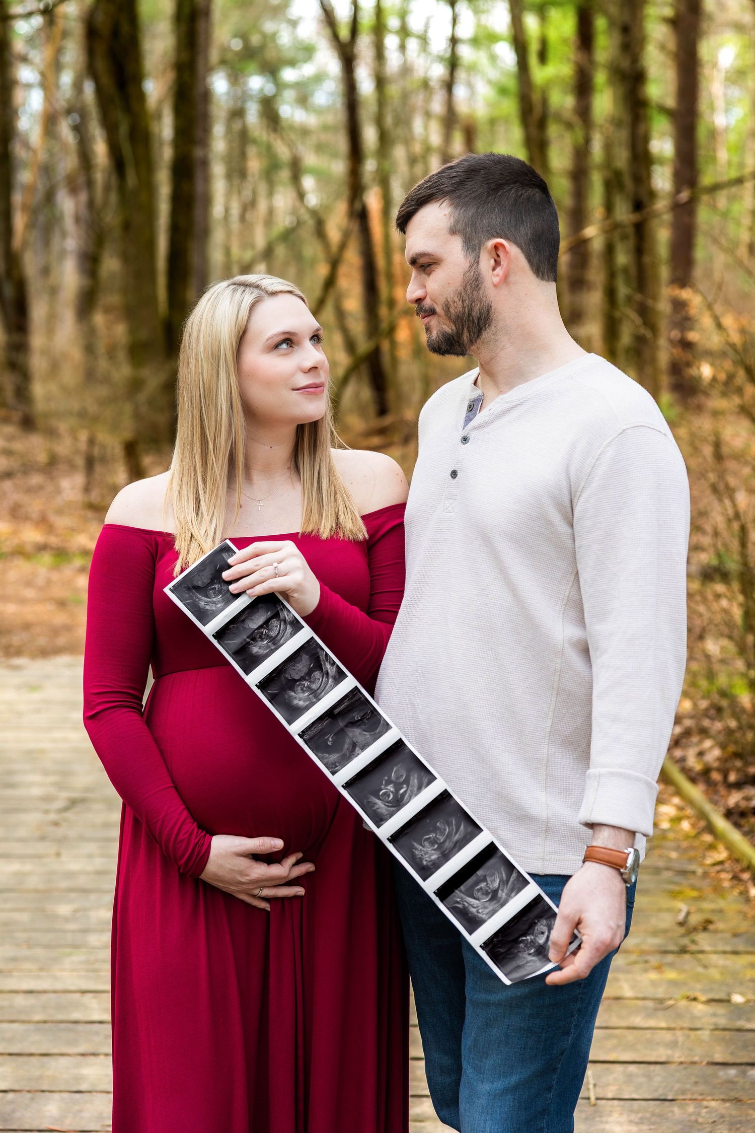 A couple stands together on a wooden boardwalk in a forest setting while holding ultrasound photos.