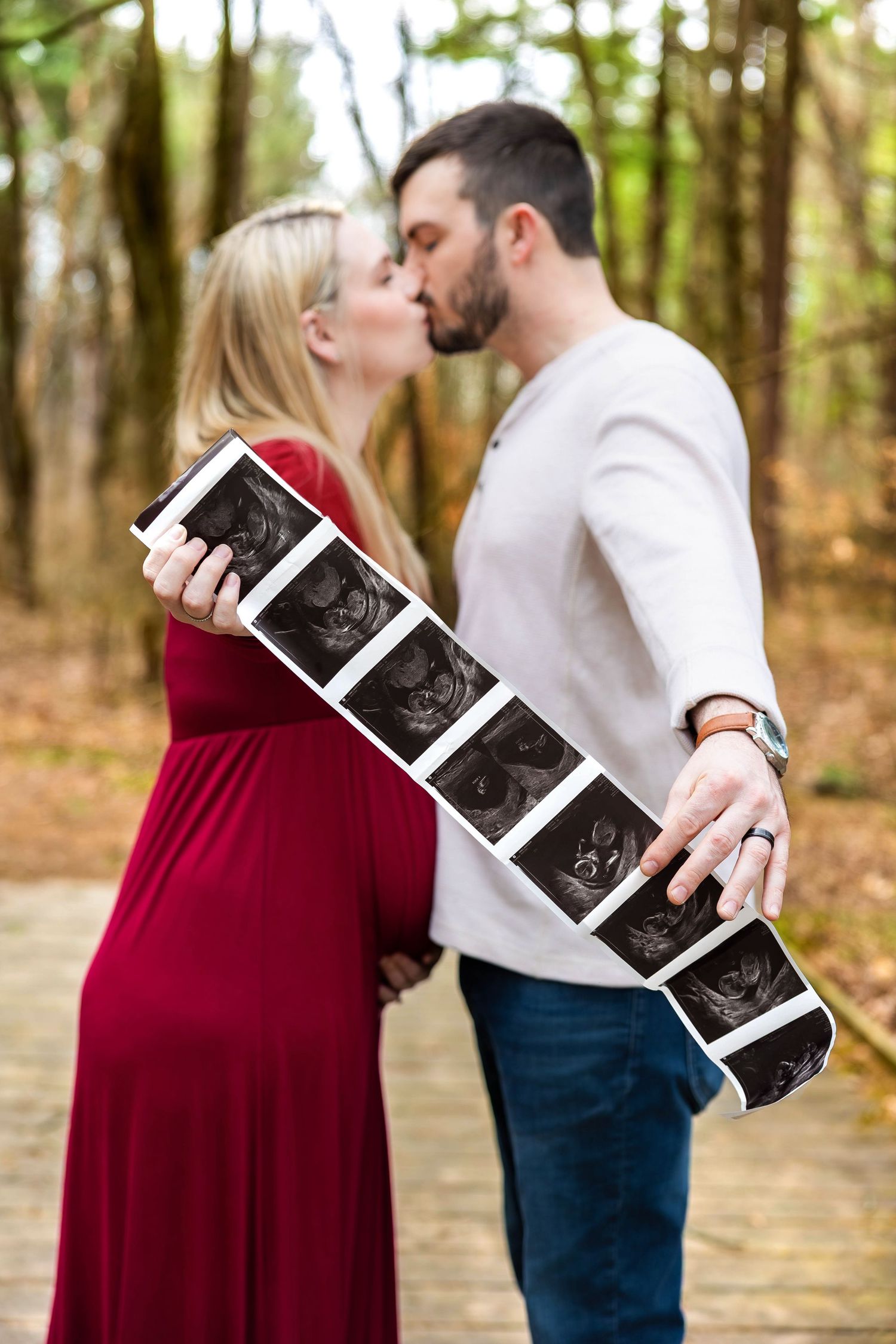 A couple shares a romantic moment in the woods while holding ultrasound photos during a pregnancy announcement photoshoot.