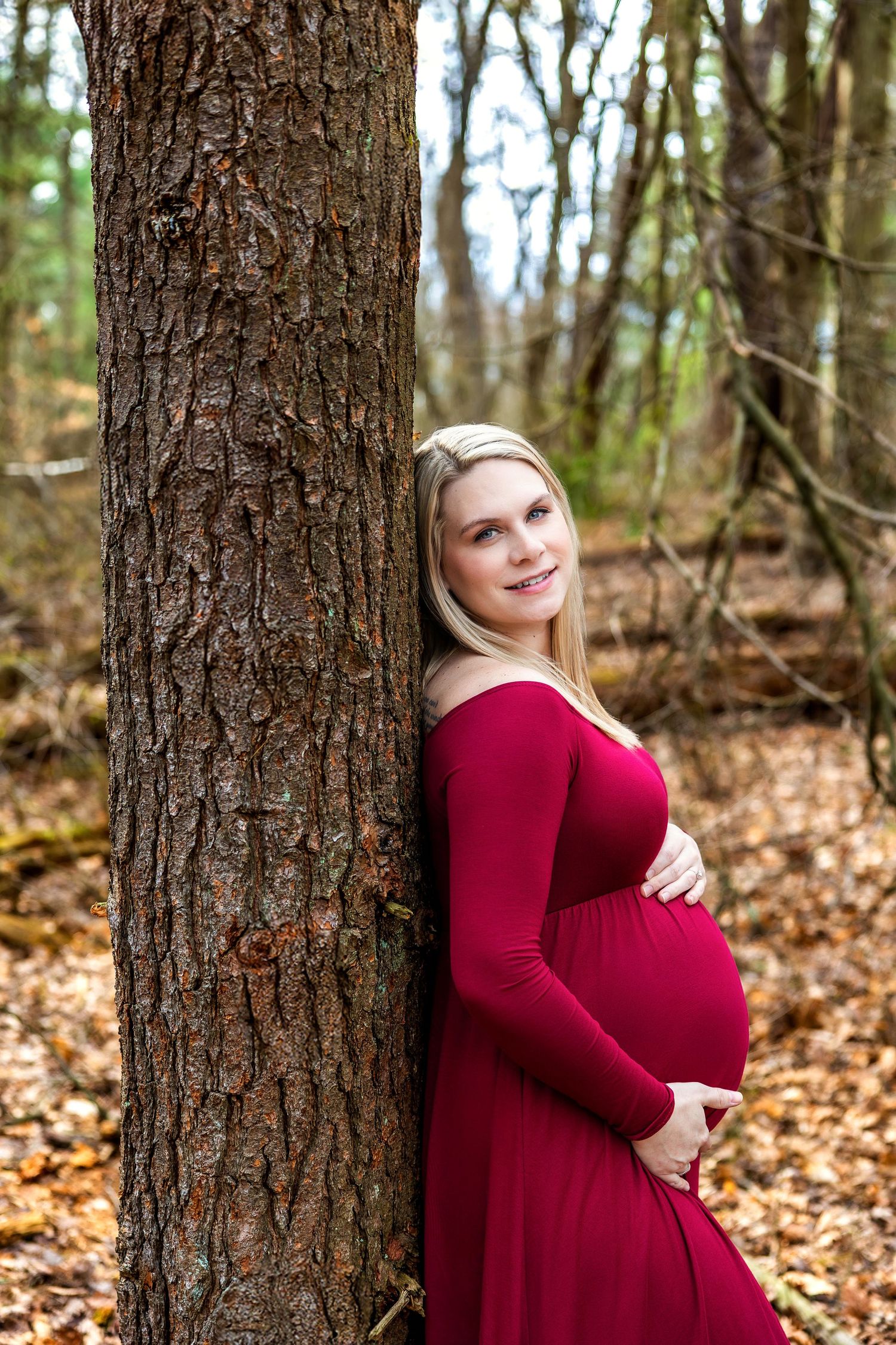 A pregnant woman in a burgundy dress poses against a tree trunk in an autumn forest setting with fallen leaves.