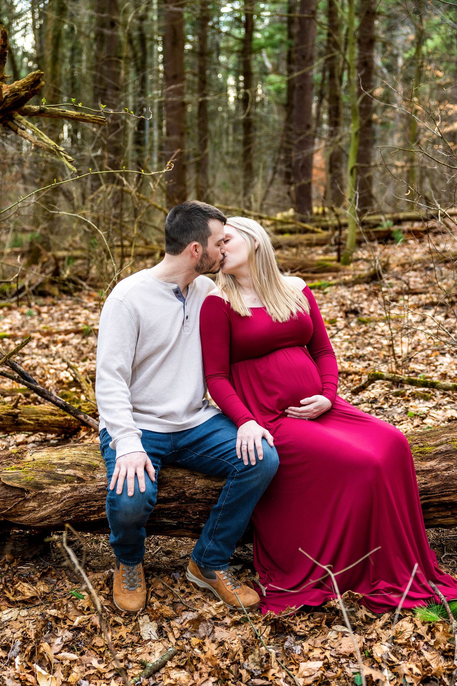 A couple in a red dress and white shirt share intimate moments while sitting on fallen logs in an autumn forest.