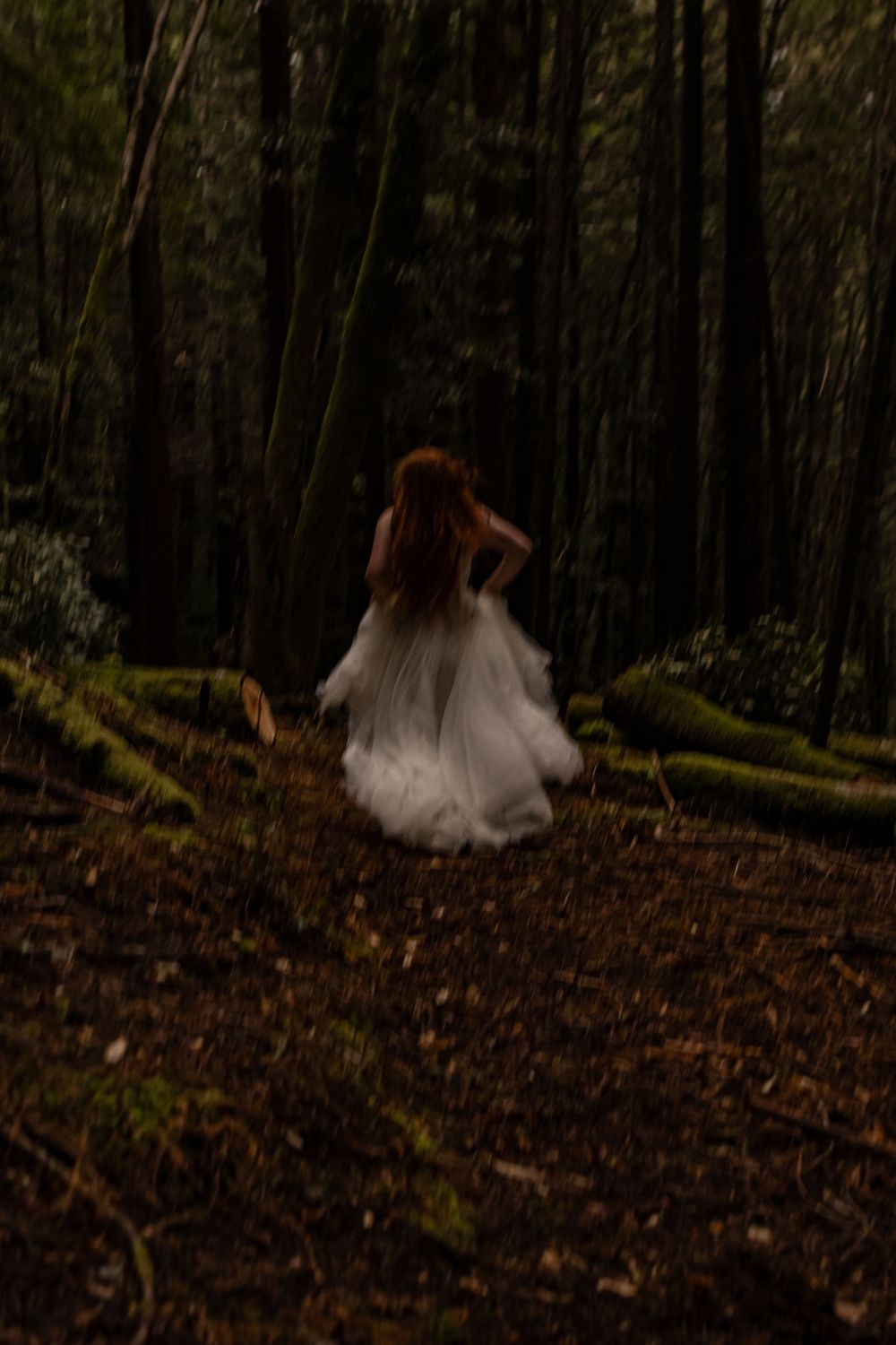A redheaded model run away from the camera in her wedding dress in the redwoods