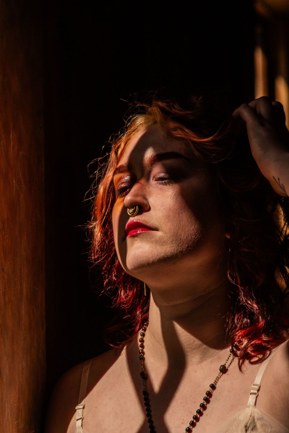 A nonbinary model is posing in the school house against a window. They're wearing red lips and you can see spaghetti straps and a necklace