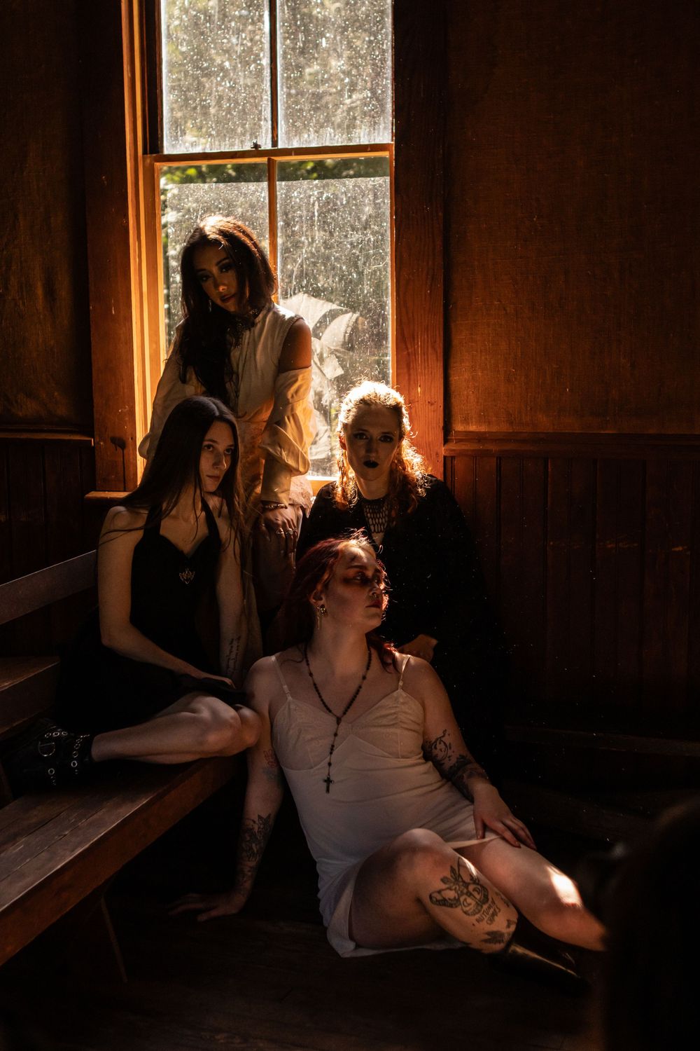 A group of four people in dresses sit in an old school house with sunlight streaming through the window
