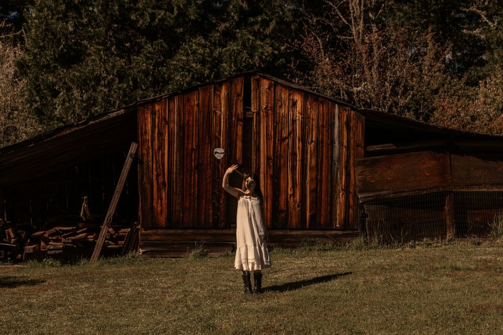 Young dark haired model poses in front of a barn in the sunlight. She's wearing some vintage lace and black boots.