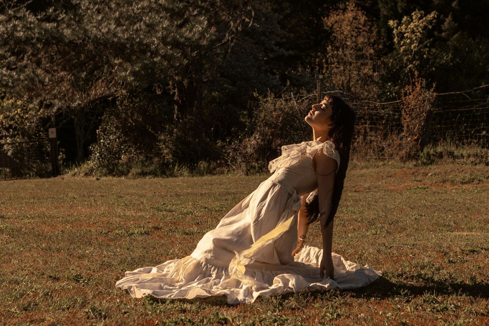 A young woman leans back in the sunlight, her profile is backlit. She has dark hair and is wearing a vintage lace wedding dress.
